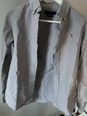 Grå skjorta från Ralph Lauren S - Klassisk grå skjorta från Ralph Lauren i custom fit med button-down krage och broderad logga på bröstet. Skjortan har långa ärmar och stängs med vita knappar framtill. Perfekt till jeans eller chinos.