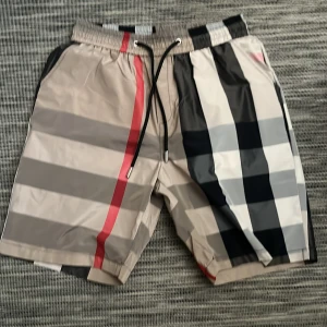 Rutig badshorts med snörning - Snygga badshorts med klassiskt rutigt mönster i beige, svart, vitt och grått samt en röd detalj. Shortsen har snörning i midjan och sidofickor. Perfekta för stranden eller poolen.