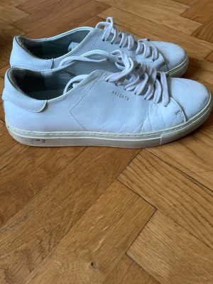 Vita sneakers från Axel Arigato - Säljer ett par klassiska vita sneakers från Axel Arigato. Skorna har rund tå, snörning och är tillverkade i slätt skinn. Minimalistisk design med diskret logga i guld på sidan. Passar perfekt till jeans eller shorts för en clean look.