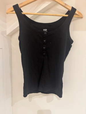 Svart ribbad linnetopp med knappar - Säljer en svart ribbad linnetopp från 157 med breda axelband och fyra knappar framtill. Toppen är ärmlös och har en enkel, stilren design som passar perfekt till jeans eller kjol. Materialet känns mjukt och stretchigt.