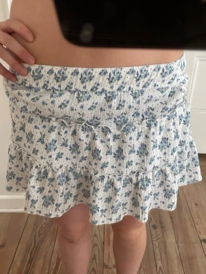 Blommig volangkjol med shorts - Supersöt kort kjol med volanger och inbyggda shorts. Vit botten med blått blommönster och tunna vertikala ränder. Kjolen har elastisk midja och är gjord i ett mjukt, stretchigt material som känns bekvämt och luftigt.