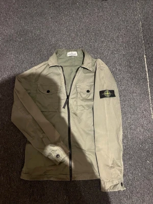 Grön overshirt från Stone Island - Säljer en grön overshirt från Stone Island med klassisk patch på ärmen. Jackan har två bröstfickor med knappar, dragkedja framtill och en avslappnad krage. Tillverkad i bomull och perfekt för lager-på-lager-stil.