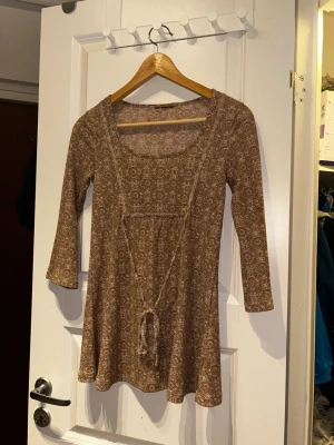 Brun mönstrad blus med knyt - Blus  i brun nyans med diskret blommigt mönster och trekvartsärm. Klänningen har en markerad midja med knytband framtill och en mjuk, stretchig passform. Perfekt för dig som gillar boho-vibbar och vill ha något bekvämt men ändå snyggt.