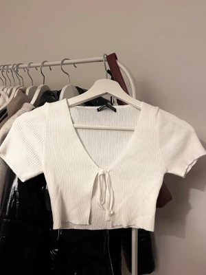 Vit ribbad croppad topp från SHEIN - Säljer en vit ribbad croppad topp från SHEIN i storlek S. Toppen har korta ärmar, djup v-ringning och en söt knytning framtill. Perfekt till jeans eller kjol för en trendig look. Materialet är mjukt och stretchigt för skön passform.