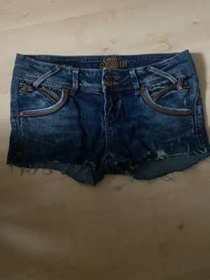 Blå jeansshorts från LTB, storlek 25 - Säljer ett par blå jeansshorts från LTB i storlek 25. Shortsen har råa, fransiga benslut, dubbla knappar i midjan och coola detaljer som nitar och dragkedjor vid fickorna. Klassisk femficksmodell med kontrastsömmar och slitningar för en avslappnad look.
