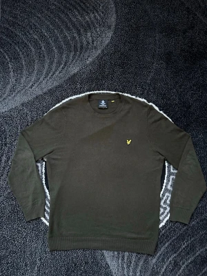 Mörkgrön stickad tröja Lyle & Scott - Snygg mörkgrön stickad tröja från Lyle & Scott med klassisk rund hals och den ikoniska gula logon broderad på bröstet. Tröjan har ribbade muddar vid ärmslut och nederkant, perfekt för lager på lager under höst och vinter.