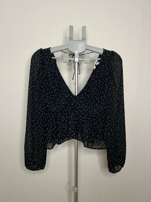 Svart prickig blus från H&M - Supersöt svart blus från H&M med vita prickar, v-ringning och långa ärmar. Modellen är croppad med volang nertill och har knytning i nacken. Tunt och luftigt material som passar perfekt till jeans eller kjol. Använd en gång.. 