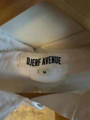 Vit skjorta från Djerf Avenue M - Klassisk vit skjorta från Djerf Avenue i storlek M. Skjortan har en bröstficka med diskret broderad logga och knappar framtill. Tillverkad i ett mjukt bomullstyg och har lång ärm och avslappnad passform.