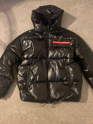 Prada jacka  - Säljer en svart pufferjacka från Prada med glansig finish och huva. Jackan har en röd logodetalj på bröstet, två sidofickor med dragkedja och snörning nedtill. VI KAN FÖRHANDLA PRISET PÅ DITT VILKOR!!!!