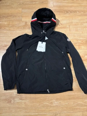 Svart Moncler vindjacka med huva - Säljer en svart vindjacka från Moncler med huva och snygga rödvita detaljer på kanten. Jackan har dragkedja framtill, två fickor med tryckknappar och Moncler-logga på ärmen. Tillverkad i lätt polyester, perfekt för blåsiga dagar.