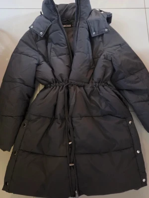 Svart lång pufferjacka från Pieces - Säljer en grå lång pufferjacka från Pieces med hög krage och justerbar midja med snörning. Jackan har dragkedja framtill, tryckknappar längs sidorna och är quiltad för extra värme. Perfekt för kalla dagar och har en clean, modern look. Är endast testad.