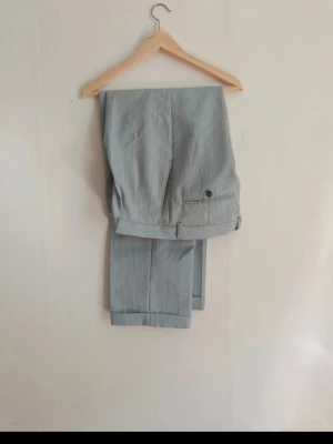 Ljusgrå kostymbyxor med pressveck - Snygga ljusgrå kostymbyxor med diskreta ränder och klassiska pressveck. Byxorna har hällor för bälte, bakficka med knapp och uppvikta benslut. Perfekta för en stilren look.