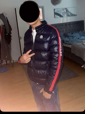 Moncler mörkblå pufferjacka - Säljer en mörkblå pufferjacka från Moncler med röd insida och bred röd rand längs ärmen där Moncler-loggan är broderad i vitt. Jackan har dragkedja framtill, ståkrage och två fickor med knappstängning. Perfekt för kalla dagar.