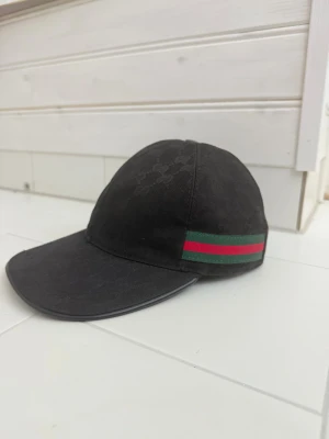Gucci canvas baseball keps - Säljer en svart keps från Gucci med diskret GG-mönster i tyget och klassiska gröna och röda ränder på sidan. Kepsen har justerbart spänne i läder baktill och är tillverkad i bomull. Perfekt accessoar för en trendig look.