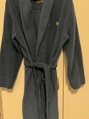 Mörkblå morgonrock från Lyle & Scott - Mysig mörkblå morgonrock från Lyle & Scott med broderad gul logga på bröstet. Västen har två stora fickor framtill och knytskärp i midjan för extra komfort. Perfekt för chill dagar hemma eller när du vill ha något riktigt mjukt på dig.