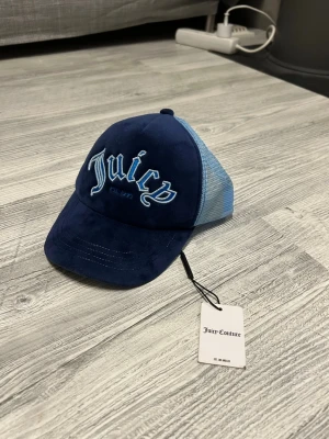 Blå meshkeps Juicy Couture - Säljer en snygg blå meshkeps från Juicy Couture med broderad text framtill. Kepsen har mörkblå front i mjukt material och ljusblå mesh baktill samt justerbar snapback. Perfekt för en cool och avslappnad stil.