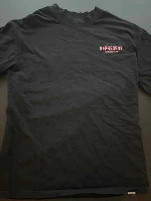 Represent svart t-shirt - Svart t-shirt från Represent med rosa tryck fram och stort tryck bak där det står 'REPRESENT OWNERS' CLUB'. Klassisk passform och rund hals. Tillverkad i mjuk bomull som känns skön mot huden. Perfekt för dig som gillar streetwear och vill sticka ut. (Storlek är S men passar mer som M)