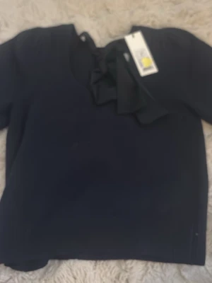 Mörkblå topp med rosett från Neo Noir - Supersnygg mörkblå topp från Neo Noir med en stor rosett vid halsen. Modellen är enkel men har en cool detalj framtill som gör den extra speciell. Perfekt till jeans eller kjol för en trendig look.