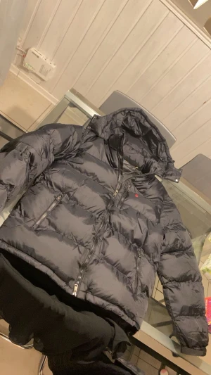 Svart pufferjacka från Polo Ralph Lauren - Säljer en svart pufferjacka från Polo Ralph Lauren med huva och dragkedja framtill. Jackan har två sidofickor med dragkedja och en liten röd logga på bröstet. Perfekt för kalla dagar och riktigt skön att bära. Pris går att diskutera 