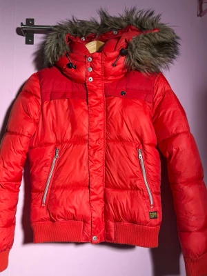Röd pufferjacka med fuskpäls från G-Star - Säljer en röd pufferjacka från G-Star med huva och fuskpäls. Jackan har dragkedjor på fickorna, ribbade muddar och snygga detaljer med knappar och snörning vid halsen. Perfekt för kalla dagar när du vill ha både stil och värme.