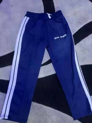 Blå Palm Angels trackpants med vita ränder - Säljer ett par blå trackpants från Palm Angels med vita och svarta ränder längs sidorna. Byxorna har resårmidja med vit snörning och Palm Angels-logga broderad på ena benet. Tillverkade i ett glansigt syntetmaterial för en sportig vibe. Priset kan diskuteras. Kommer med Palm angel posen 