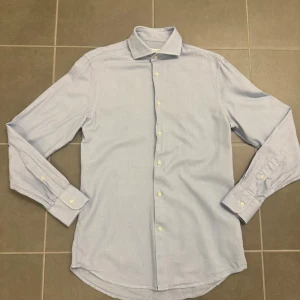 Ljusblå skjorta från J.Lindeberg - Säljer en stilren ljusblå skjorta från J.Lindeberg i storlek 38. Skjortan har klassisk krage, långa ärmar och vita knappar framtill. Materialet är mjukt och känns som bomull, perfekt till många olika looks.