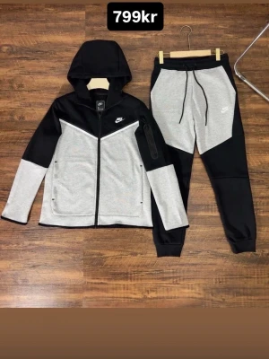 Grå och svart Nike väst - Snygg väst från Nike i grått och svart med dragkedja framtill och vit logga på bröstet. Västen har en sportig look med kontrastpaneler och är tillverkad i mjukt material som känns bekvämt. Perfekt för dig som gillar streetwear och sportig stil.