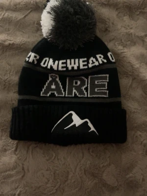 Svart och vit Åre-mössa med tofs - Säljer en svart och vit mössa från Onewear med stor tofs på toppen och texten 'ÅRE' framtill. Mössan har ett bergmotiv på uppvikt kant och stickat mönster runt om. Perfekt för kalla dagar och vintersäsongen.