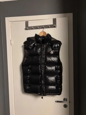 Svart dunväst från Moncler - Säljer en svart, glansig dunväst från Moncler med avtagbar luva och dragkedja framtill. Västen har Moncler-logga på bröstet och två fickor med dragkedja. Storlek 2. Fungerande NFC-scan