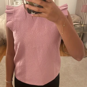 Rosa ärmlös topp med volang från Pieces - Andvänt 3 gångerSupersöt ljusrosa topp från Pieces med strukturerat tyg och volangdetaljer på axlarna. Toppen är ärmlös och har rund halsringning. Perfekt för dig som gillar en feminin och trendig look. Parfekt topp till sommaren.