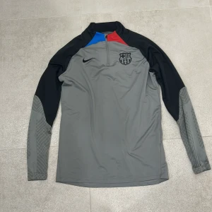 FC Barcelona långärmad träningströja Nike - Snygg långärmad träningströja från Nike med FC Barcelona-logga på bröstet. Tröjan är grå med svarta ärmar och har detaljer i blått och rött vid kragen. Tillverkad i ett lätt och funktionellt material, perfekt för fotbollsträning.