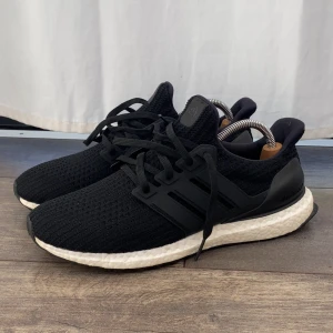 Adidas Ultraboost svarta sneakers - Svarta Adidas Ultraboost sneakers med stickad ovandel i syntet och klassiska tre ränder på sidan. Skorna har vit, responsiv Boost-sula och svart yttersula. Perfekta för dig som vill ha både stil och komfort. Snörning framtill och diskret logga på hälen.