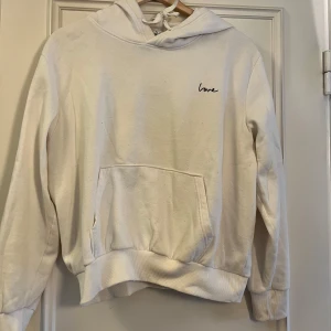 Vit hoodie med broderad text  - Vit hoodie med broderad svart text 'love' på bröstet. Klassisk känguruficka framtill, ribbade muddar och huva med snörning. Mjuk och skön, perfekt för chill dagar. Enkel och stilren design som passar till det mesta.