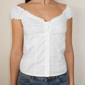 Vit blus med volang och knappar - Söt vit blus med korta puffärmar och volangkant längs den djupa v-ringningen. Blusen har knäppning framtill och är figursydd för en feminin siluett. Perfekt till jeans eller kjol för en fräsch look.