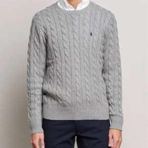 Grå kabelstickad tröja från Ralph Lauren - Kabelstickad grå tröja från Ralph Lauren med klassisk rund halsringning och diskret broderad logga på bröstet. Tröjan har långa ärmar och ribbade muddar, perfekt för lager-på-lager-stil under kyliga dagar.