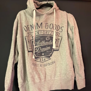 Grå hoodie från Smog med tryck - Säljer en grå hoodie från Smog med stort svart tryck på bröstet där det står 'Denim Goods Authentic Denim'. Tröjan har huva med vita snören och är i regular fit. Perfekt för chill dagar eller när du vill ha en avslappnad stil.