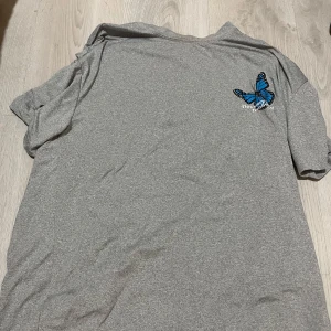 Grå t-shirt med blå fjärilstryck - Snygg grå t-shirt med stort blått fjärilstryck och texten 'This story is not finished' på ryggen. T-shirten har korta ärmar och är tillverkad i mjukt material, perfekt för en chill och avslappnad stil.