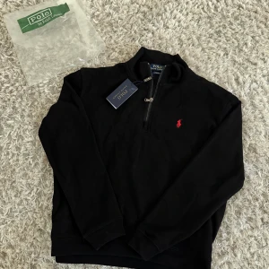 Svart half zip tröja Polo Ralph Lauren - Svart långärmad tröja från Polo Ralph Lauren med röd broderad logga på bröstet. Tröjan har en snygg half zip-dragkedja och ribbade muddar. Klassisk och stilren modell i bomull, perfekt för lager-på-lager.