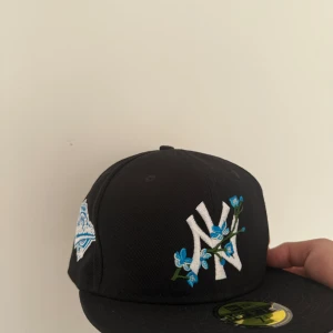 New Era NY keps med blå blommor - Snygg svart New Era 59FIFTY keps med vit NY-logga och blå blommor broderade framtill. På sidan finns ett broderat World Series 1996-märke i blått och vitt. Kepsen har rak skärm och klassisk passform.