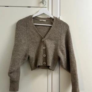 Beige croppad kofta från Gina Tricot - Superfin beige croppad kofta från Gina Tricot i ribbstickad ullmix. Koftan har v-ringning, långa ärmar och stora knappar framtill. Perfekt för lager-på-lager och ger en chill men trendig vibe till din outfit.