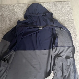 Under Armour vindjacka blå/grå - Snygg vindjacka från Under Armour i marinblått och grått. Jackan har huva, hel dragkedja, bröstficka med dragkedja och elastiska muddar med logga. Perfekt för blåsiga dagar och sportiga outfits.