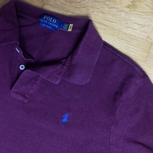 Vinröd Polo Ralph Lauren pikétröja - Säljer en vinröd pikétröja från Polo Ralph Lauren i klassisk passform. Tröjan har korta ärmar, krage och knäppning framtill samt den ikoniska blå broderade loggan på bröstet. Tillverkad i mjuk bomull för en skön känsla.