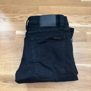Svarta jeans från Nudie Jeans - Säljer ett par svarta jeans från Nudie Jeans med klassisk femficksdesign och diskret logga på bakfickan. Jeansen har raka ben och normal passform, tillverkade i mjuk bomull med en stilren look. Perfekta för dig som gillar minimalistisk stil.