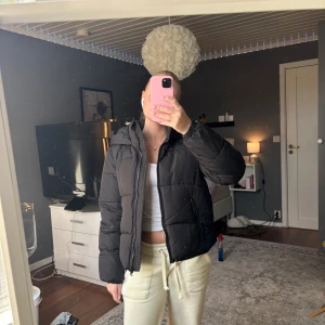 Svart pufferjacka med huva - Säljer en svart pufferjacka med huva och dragkedja framtill. Jackan har quiltad design, två fickor med dragkedja och är långärmad. Perfekt för kyligare dagar och riktigt skön att bära. Passformen är normal och modellen är klassisk och enkel att matcha.