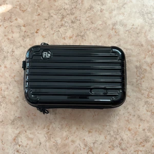 Väska  - Snygg svart liten resväska från FL med glansig yta och räfflat mönster. Perfekt för att förvara småprylar eller använda som necessär. Väskan har dragkedja runtom och meshfickor på insidan för extra organisering.