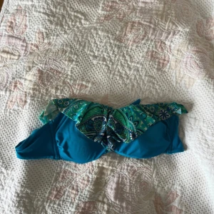 Turkos bikiniöverdel från Tezenis - Snygg turkos bikiniöverdel från Tezenis med paisley-inspirerat mönster i grönt och blått på volangen. Bandeau-modell med avtagbar axelrem i samma mönster. Perfekt för strandhäng och poolparty! Materialet är mjuk och stretchig polyester.