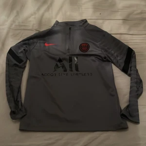 PSG långärmad träningströja Nike - Snygg grå långärmad fotbollströja från Nike med PSG-logga på bröstet och röd Nike swoosh. Tröjan har halv dragkedja och är tillverkad i Dri-FIT-material som andas. Perfekt för träning eller när du vill representera Paris Saint-Germain.