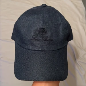 Mörkblå keps från Loro Piana - Snygg mörkblå keps från Loro Piana med broderad logga framtill. Kepsen har justerbar rem med metallspänne bak och insida i rutigt tyg. Tillverkad i exklusivt Storm System-material som är både stilrent och funktionellt. Perfekt för dig som gillar premiumaccessoarer.