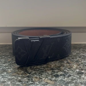 Svart bälte  - Snyggt svart  bälte i skinn med klassiskt monogrammönster och i metall. Bältet har en brun insida. Perfekt accessoar för att lyfta din outfit med en lyxig touch.