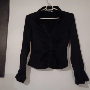 Svart skjorta med knappar från Zara - Svart skjorta från Zara med snygg rynkdetalj framtill och klassisk krage. Skjortan har långa ärmar och knäppning med fyra knappar. Materialet är glansigt och känns mjukt mot huden, vilket ger en elegant vibe.